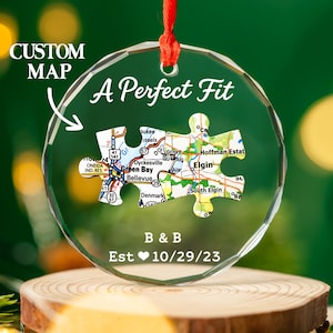 Pode incluir: Um ornamento circular de vidro transparente com uma fita vermelha. No centro, duas peças de puzzle entrelaçadas exibem um mapa de Green Bay e Elgin. O texto diz A Perfect Fit, B & B e Est 10/29/23. O vidro tem uma borda facetada.