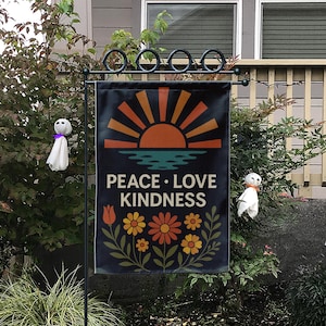 Könnte beinhalten: Eine schwarze Gartenflagge mit einem Retro-Sonnenuntergangsdesign, den Worten "PEACE LOVE KINDNESS" und floralen Akzenten. Die Flagge hängt an einem schwarzen Metallständer, mit zwei kleinen Geisterdekorationen.