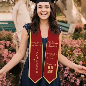 May include: A smiling graduate wearing a black cap and a dark blue dress. She is wearing a red and gold sash that says "University of Michigan Engineer" and "Clara Smith Bachelor of Science Class of 2026".