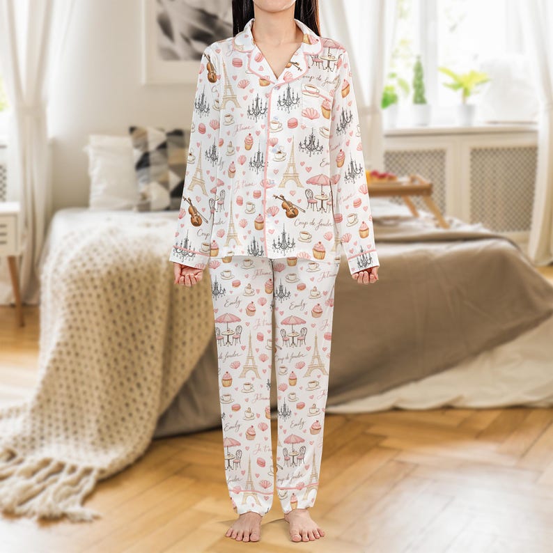 May include: White pajama set with a repeating pattern of the Eiffel Tower, chandeliers, musical instruments, and cafe scenes in pink and beige. The set includes a button-down top and pants, trimmed with pink piping.