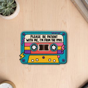 Könnte beinhalten: Ein rechteckiger, bestickter Aufnäher in Form einer Kassette mit türkisfarbenem Rand. In der Mitte steht der Text "PLEASE BE PATIENT WITH ME, I'M FROM THE 1990S." Das Design enthält rosa, gelbe und violette Akzente mit Blitzmotiven.