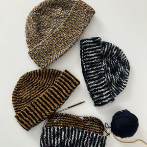 May include: Four knitted beanies in various color combinations. One is in progress with knitting needles and stitch markers. Colors include brown, black, white, and gold. A ball of dark blue yarn is also present.