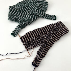 May include: Two knitted items, one in green and white stripes, the other in black and tan stripes. The green and white item is folded. The black and tan item is partially knitted, with needles and yarn visible.