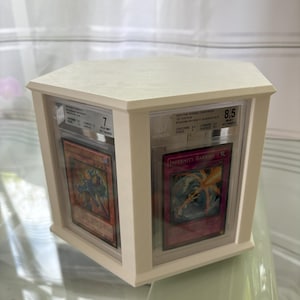 Könnte beinhalten: Ein weißer sechseckiger Display-Kasten mit drei Fächern für Sammelkarten. Der Kasten hat eine transparente Acrylglasfront und einen weißen Hintergrund. Die Karten sind durch das Acrylglas sichtbar. Die Karten sind bewertet und haben ein weißes Etikett mit schwarzem Text. Der Text auf dem Etikett enthält den Kartennamen, die Edition, die Bewertung und das Bewertungsunternehmen. Die Karten sind "Drive the Banking Darkness" und "Infinity Barrier".