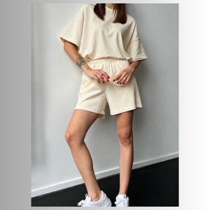 May include: A cream-colored two-piece outfit featuring a short-sleeved t-shirt and matching shorts. The shorts have an elastic waistband and a relaxed fit.