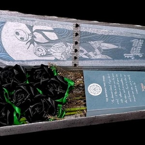 May include: A coffin-shaped box with a hinged lid, featuring a black and white illustration of Jack Skellington and Sally. Inside are black ribbon roses with green accents, a bouquet of dried flowers, and a book with the title "The Nightmare Before Christmas".