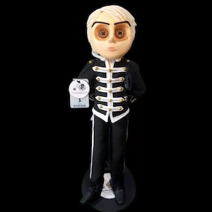 Könnte beinhalten: Handgefertigte Figur einer Person in einer schwarzen Uniform im Militärstil mit weißen Verzierungen und goldenen Knöpfen. Die Figur hat einen hellen Teint, blondes Haar und Knopfaugen. Sie hält ein Etikett mit einem Oktopus-Design und dem Text "THE BLACK PARADE".