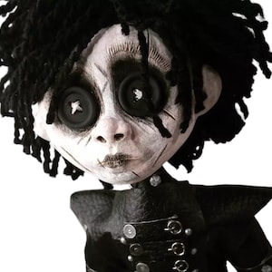 May include: A handcrafted doll with a pale face, black button eyes, and stitched mouth. The doll has black yarn hair and wears a black outfit with silver-colored accents. The doll's face has dark eye makeup and painted scars.