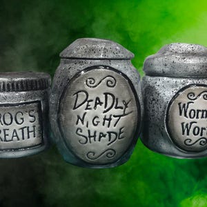 Può includere: Tre barattoli grigi strutturati con coperchi, etichettati "Frog's Breath", "Deadly Nightshade" e "Worm's Wort". I barattoli hanno un aspetto invecchiato, che suggerisce un tema di pozioni o di farmacia.