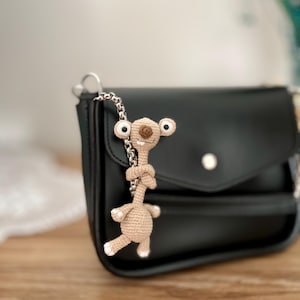 Puede incluir: Un pequeño personaje beige tejido a ganchillo con ojos grandes y redondos y una cadena está unido a un bolso negro. El personaje tiene detalles blancos en los pies y una nariz marrón. El bolso tiene una correa de cadena plateada y un cierre plateado.