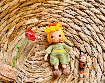 May include: Crocheted doll of a character in a green outfit with a yellow belt and hair, and a small fox. A glass bottle with a red flower is also present. The items are on a woven surface.