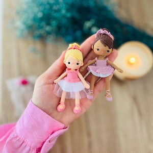 Puede incluir: Dos muñecas de bailarina de ganchillo hechas a mano. Una muñeca tiene cabello rubio y un tutú rosa, la otra tiene cabello castaño y un tutú rosa. Ambas muñecas se sostienen en una mano, con un fondo borroso de flores azules y una vela.