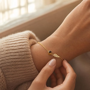 Op de afbeelding: Een goudkleurige kettingarmband met een kleine, ronde bedel met een donker midden en een delicaat, opengewerkt vleugelbedeltje aan de zijkant. De armband is gemaakt van een fijne schakelketting en sluit om de pols.