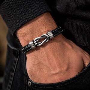 Könnte beinhalten: Ein schwarzes geflochtenes Lederarmband mit einem zentralen Knotenornament aus silberfarbenem Metall. Der Knoten wird von zwei Sätzen aus drei silberfarbenen Metallringen flankiert, die das Ornament mit dem schwarzen Lederband verbinden.