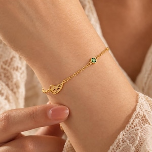 Könnte beinhalten: Ein goldfarbenes Kettenarmband mit einem kleinen, zierlichen Flügelanhänger und einem einzelnen runden, grün gefassten Edelstein. Das Armband besteht aus feinen Gliedern und verfügt über einen Verschluss, was ein schlichtes Design ergibt.