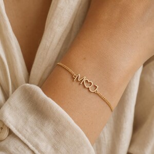 Op de afbeelding: Een goudkleurige kettingarmband met een centrale metalen bedel met de letters M en J, gescheiden door een kleine hartvorm. De fijne schakelketting is verbonden met het centrale letterstuk, wat zorgt voor een doorlopend sieraad.