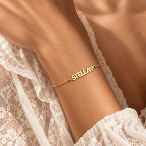 Op de afbeelding: Een goudkleurige kettingarmband met de naam STELLA in hoofdletters, gevolgd door een kleine hartvormige bedel ingelegd met heldere steentjes. De armband is bevestigd met een fijne schakelketting die om de pols zit.