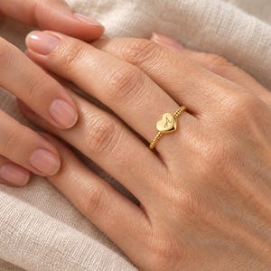 Op de afbeelding: Een goudkleurige ring met een kleine hartvormige bedel gegraveerd met de letter A. De ringband heeft een delicate kralentextuur, wat zorgt voor een consistente metallic afwerking over het hele sieraad dat om een vinger wordt gedragen.