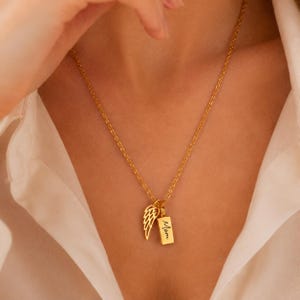 Pode incluir: Um colar de tom dourado com uma corrente delicada e dois pingentes: uma asa de anjo recortada e um pequeno pendente retangular gravado com a palavra Mom em letra cursiva. A joia repousa sobre uma camisa branca de colarinho.