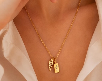 May include: A gold-toned necklace featuring a delicate chain and two charms: a cutout angel wing and a small rectangular pendant engraved with the word Mom in a cursive script. The jewelry rests against a white collared shirt.