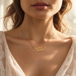 Op de afbeelding: Een goudkleurige metalen ketting met een fijne schakelketting en een gepersonaliseerde hanger. De hanger toont de namen Frederica en Jennifer in een cursief lettertype, geaccentueerd door twee kleine, open hartvormen in het ontwerp.