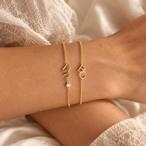 Op de afbeelding: Twee goudkleurige kettingarmbanden met verfijnde letterbedels. Eén armband toont de letters D en M met een kleine, ronde, heldere steen in de ketting. De tweede armband bevat de letters B en C in een verbonden, cursieve schrijfstijl.