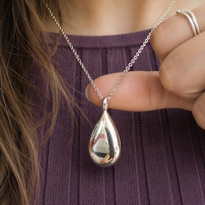 Op de afbeelding: Een gepolijste zilverkleurige druppelvormige hanger aan een fijne bijpassende ketting. Het gladde, reflecterende oppervlak van de hanger vangt het licht op en zorgt voor een heldere glans. De ketting heeft een eenvoudig en strak ontwerp.