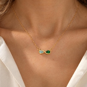 Peut inclure: Un collier à chaîne dorée avec un pendentif en forme de symbole infini. Le pendentif est orné d'une pierre bleu clair en forme de poire et d'une pierre vert émeraude en forme de poire, reliées par une fine chaîne à maillons dorés.