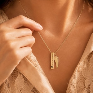 Op de afbeelding: Een goudkleurige ketting met een verticale rechthoekige staafhanger gegraveerd met de data 3.11.10 en 6.14.20 en een pootafdruk-icoon, vergezeld door een aparte goudkleurige vleugelbedel die aan een fijne ketting is bevestigd.