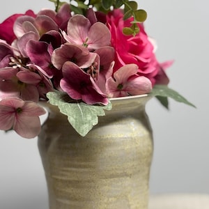 Könnte beinhalten: Eine Vase aus Keramik mit einer strukturierten, hellbraunen Oberfläche enthält ein Arrangement aus Kunstblumen. Das Arrangement umfasst tiefrosa Hortensien, eine leuchtend rosa Rose und grünes Laub.