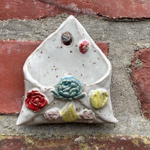 Könnte beinhalten: Eine weiße, keramikförmige Wandtasche in Umschlagform mit farbenfrohen floralen Verzierungen. Die Tasche hat eine rote Rose, eine blaue Blume und andere kleine, dekorative Elemente. Das Stück ist an einer roten Backsteinmauer befestigt.