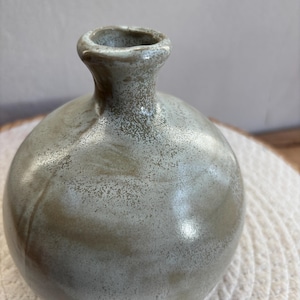 Könnte beinhalten: Eine kleine, runde Keramikvase mit schmalem Hals und bauchigem Körper. Die Vase hat eine gesprenkelte, matte Oberfläche in Grau- und Brauntönen. Die Vase steht auf einem weißen, gewebten Untersetzer.
