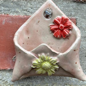 Könnte beinhalten: Eine handgefertigte, rosa Keramik-Wandtasche in Form eines Umschlags, verziert mit einer roten und einer gelben Blume. Die Tasche ist an einer Ziegelmauer befestigt und verleiht jedem Raum einen rustikalen Charme.