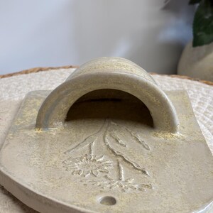 May include: A ceramic wall pocket in a light beige color, featuring a curved handle and a rectangular base. The front is decorated with an embossed floral design. The piece has a small hole at the bottom.