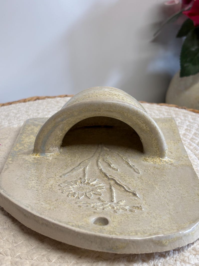 May include: A ceramic wall pocket in a light beige color, featuring a curved handle and a rectangular base. The front is decorated with an embossed floral design. The piece has a small hole at the bottom.