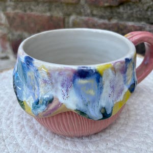 Könnte beinhalten: Ein Keramikbecher mit einer rosafarbenen Basis und vertikalen Rillenstrukturen. Der obere Teil ist mit einem marmorierten Muster aus blauen, weißen, gelben und violetten Tropfen glasiert. Das Innere ist glatt und weiß, der Henkel ist rosa.