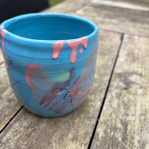 Könnte beinhalten: Eine handgefertigte Keramiktasse mit türkisfarbenem Grund und rosa Farbtropfen. Die Tasse hat abstrakte rosa und grüne Pinselstriche und ein gepunktetes Liniendesign. Die Tasse steht auf einer Holzoberfläche.