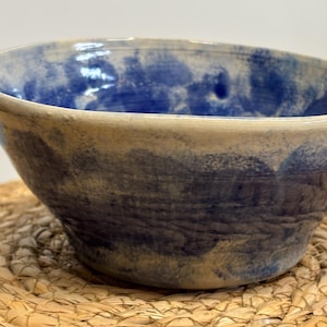 Könnte beinhalten: Eine Keramikschale mit blau-weißem, meliertem Design. Die Schale hat eine breite, flache Form und einen hellbraunen Rand. Der Innenraum weist ein dunkleres blaues Muster auf, das einen Kontrast erzeugt. Die Schale steht auf einer geflochtenen Strohmatte.