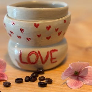 Könnte beinhalten: Ein gestapelter, cremefarbener Keramikbehälter mit roten Herzakzenten und dem Wort "LOVE" auf der Unterseite. Kaffeebohnen und rosa Blütenblätter sind auf einer Holzoberfläche um den Behälter herum verstreut.