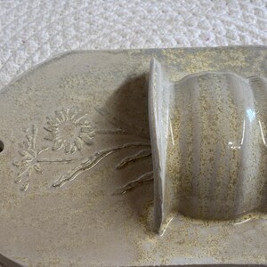 May include: A beige ceramic wall pocket with a floral design. The pocket has a rounded top with a hole for hanging and a rectangular base. The floral design is embossed on the rounded top.