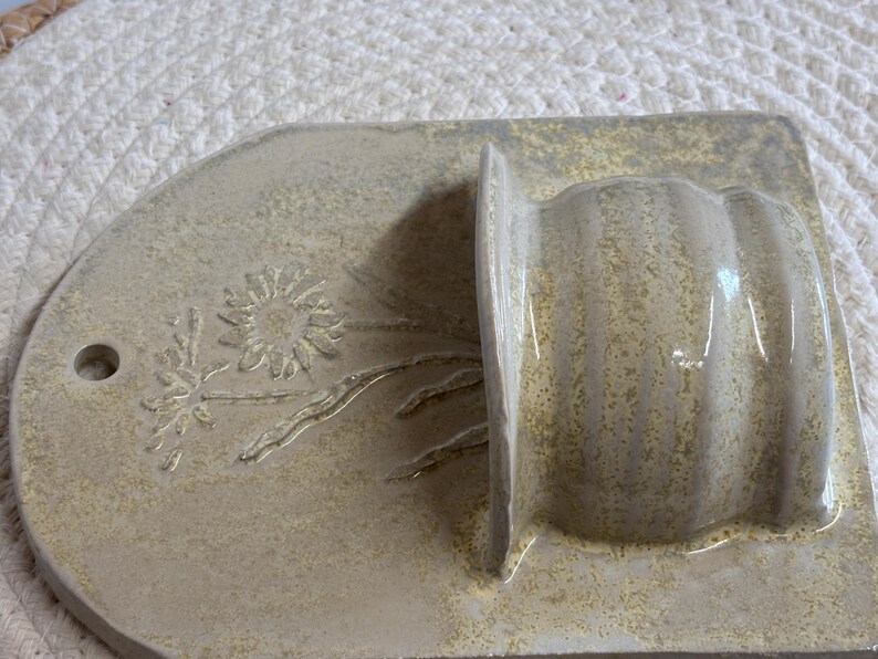 May include: A beige ceramic wall pocket with a floral design. The pocket has a rounded top with a hole for hanging and a rectangular base. The floral design is embossed on the rounded top.