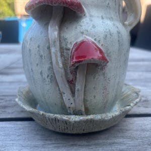 Könnte beinhalten: Ein Keramikbecher mit gesprenkelter, cremeweißer Glasur und zwei erhabenen, dreidimensionalen roten Pilzen auf der Vorderseite. Der Becher hat einen seitlichen Henkel und steht auf einer passenden, strukturierten Untertasse mit geriffeltem Rand.