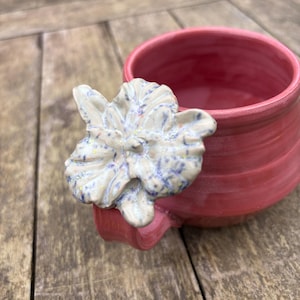 Könnte beinhalten: Eine handgefertigte, rote Keramik-Tasse mit einer weißen und blauen Blumenverzierung. Die Tasse hat eine abgerundete Form und einen kleinen Henkel. Das florale Detail verleiht der funktionalen Keramik eine dekorative Note.