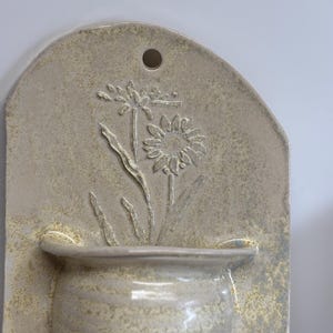 May include: A beige ceramic wall pocket with a floral design. The rectangular plaque features a raised relief of a flower and leaves above a small, rounded pot. A hole is at the top for hanging. The piece has a textured, speckled finish.