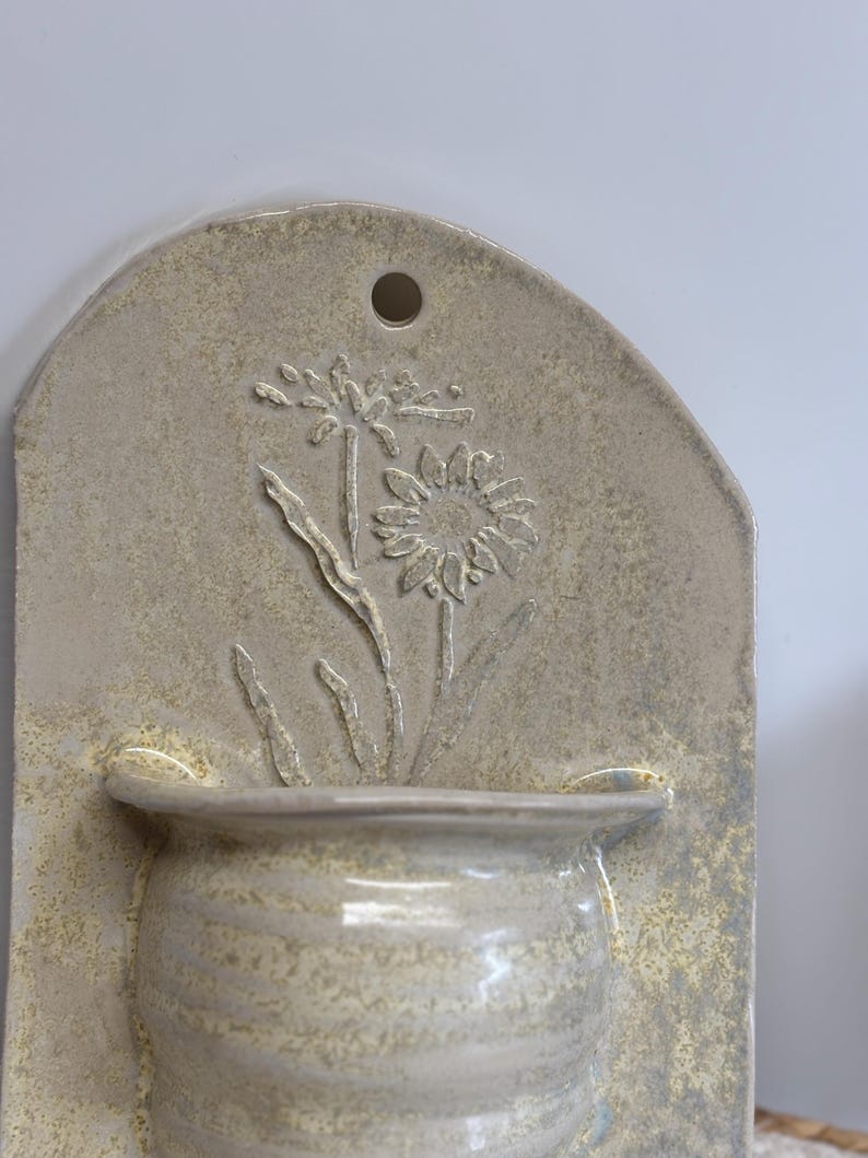 May include: A beige ceramic wall pocket with a floral design. The rectangular plaque features a raised relief of a flower and leaves above a small, rounded pot. A hole is at the top for hanging. The piece has a textured, speckled finish.