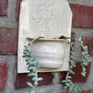 May include: A cream-colored ceramic wall planter with a floral design and a small pot for plants. The planter is mounted on a red brick wall, with trailing green succulent plants cascading from the pot.