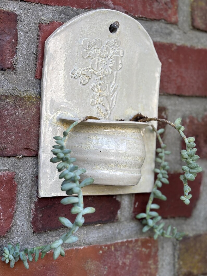 May include: A cream-colored ceramic wall planter with a floral design and a small pot for plants. The planter is mounted on a red brick wall, with trailing green succulent plants cascading from the pot.