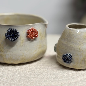 Könnte beinhalten: Zwei Keramikgefäße mit floralen Akzenten. Die größere, offene Schale hat drei Blumenverzierungen in Weiß, Marineblau und Orange. Die kleinere, runde Vase hat marineblaue und gelbe Blumendetails. Beide sind cremefarben mit einer gesprenkelten Textur.