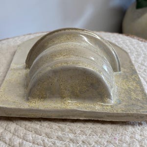 May include: A ceramic soap dish with a curved, ridged design in shades of beige and light yellow. The dish has a rectangular base and a glossy finish, suitable for holding soap bars. The overall aesthetic is minimalist and modern.