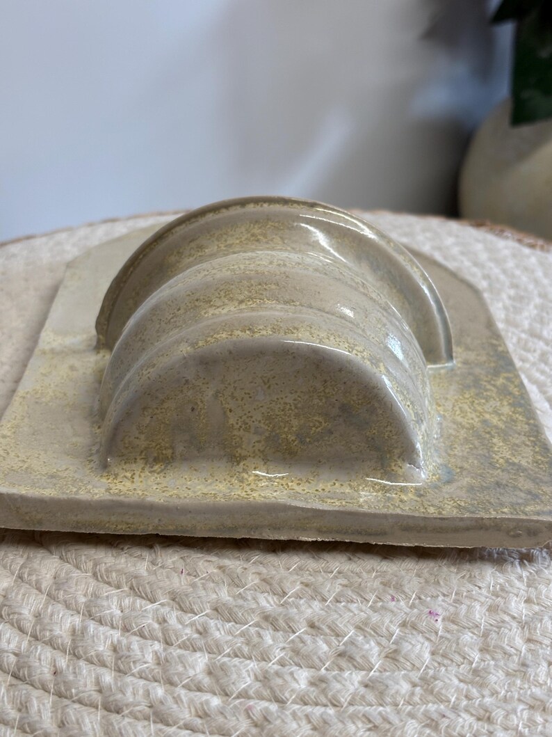 May include: A ceramic soap dish with a curved, ridged design in shades of beige and light yellow. The dish has a rectangular base and a glossy finish, suitable for holding soap bars. The overall aesthetic is minimalist and modern.